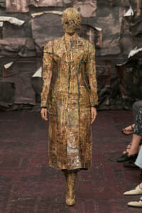 Model on a runway wearing a long, patchwork brown coat and matching boots with a textured, sculpted headpiece.
