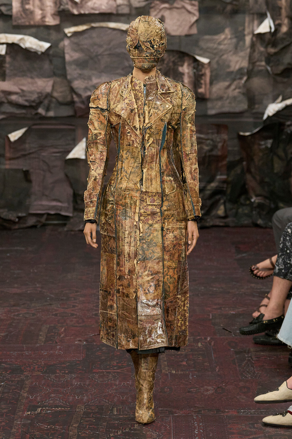 Model on a runway wearing a long, patchwork brown coat and matching boots with a textured, sculpted headpiece.