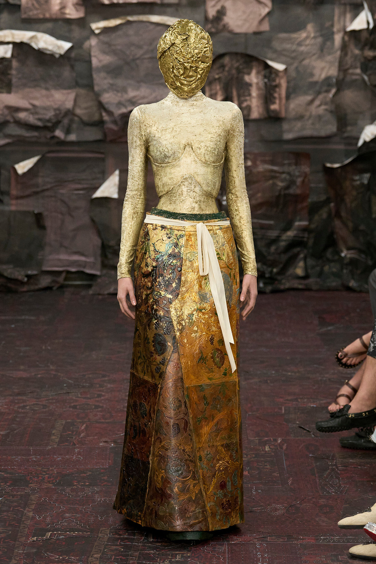 Model wearing a gold foil headwrap, cream-gold metallic top, and ornate brocade skirt with a white sash.