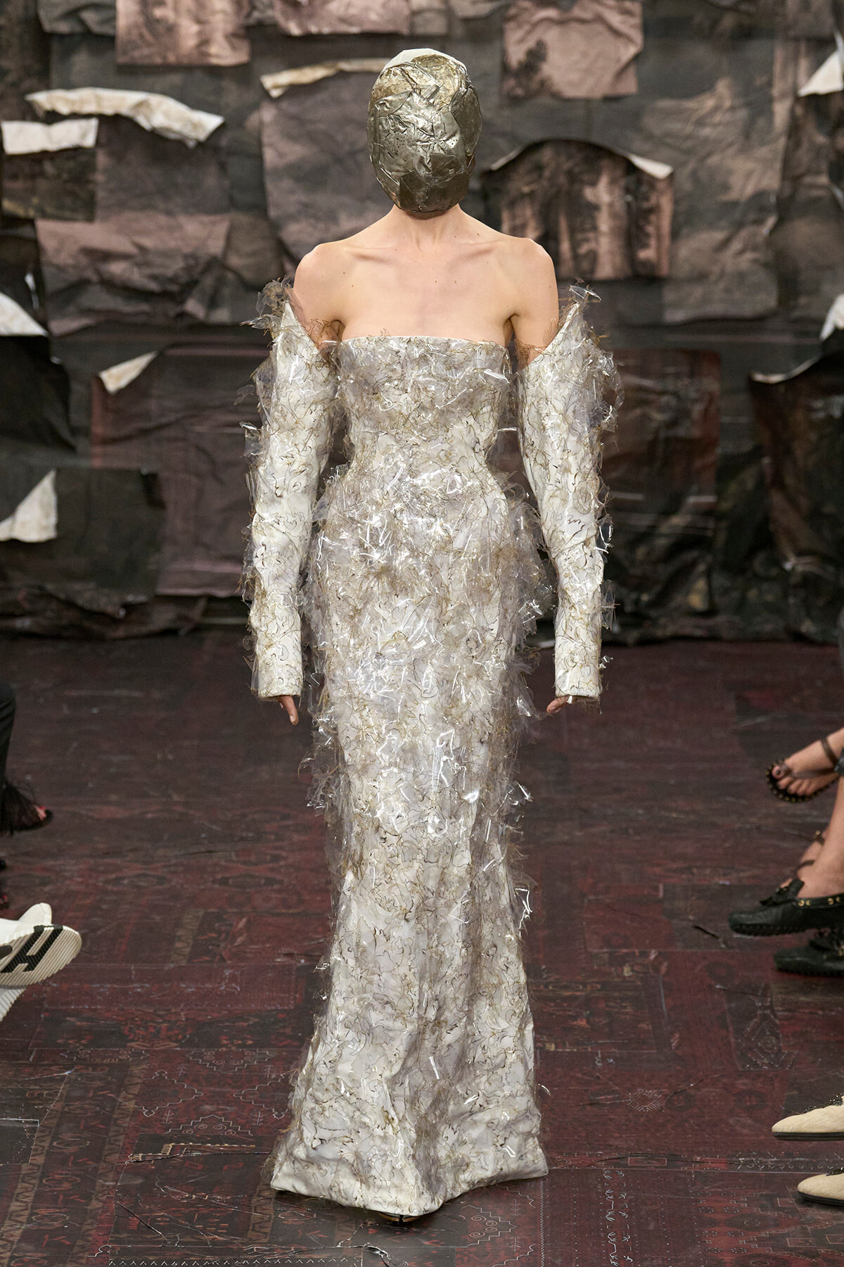 Model on runway in an off-shoulder silver gown made of shredded foil, with matching metallic mask obscuring the face.