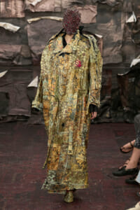 Model on a runway wearing a long, patchwork camouflage coat and a beaded red mask; hands with red nails visible.