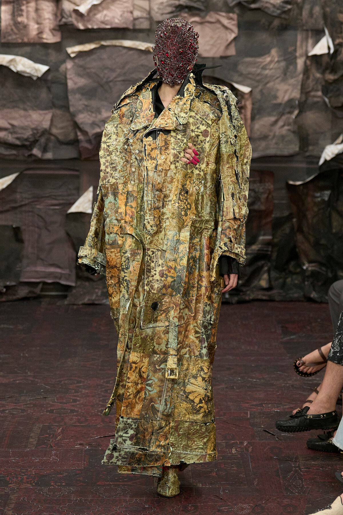 Model on a runway wearing a long, patchwork camouflage coat and a beaded red mask; hands with red nails visible.