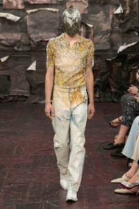 Runway model in a metallic foil headpiece and distressed, paint-splattered top with light-blue jeans, walking the catwalk.