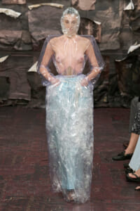 Model walks a runway in a clear plastic rain poncho over a pale outfit, head wrapped in plastic film under dim backstage lighting.