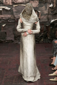 Model on a fashion runway wearing a full-length, metallic silver gown with a sculptural, beaded headpiece obscuring the face; shoulders have cut-out sleeves and the fabric has a soft sheen against a dark backdrop.
