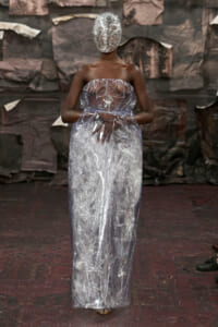 Model wearing a full-length transparent plastic gown and matching head wrap on a runway, dark background.