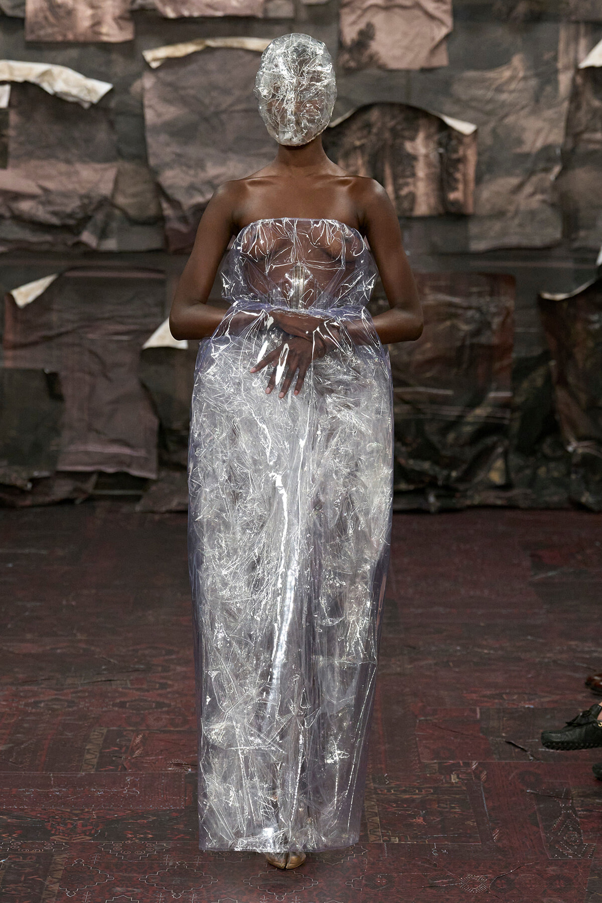 Model wearing a full-length transparent plastic gown and matching head wrap on a runway, dark background.