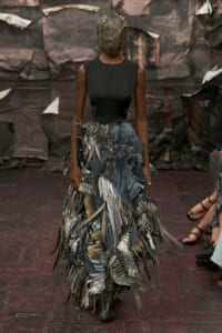 Runway model in a sleeveless black top and a voluminous feathered skirt; head covered by a metallic mask.