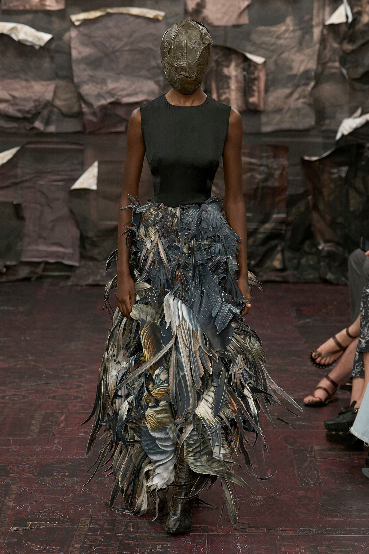 Runway model in a sleeveless black top and a voluminous feathered skirt; head covered by a metallic mask.