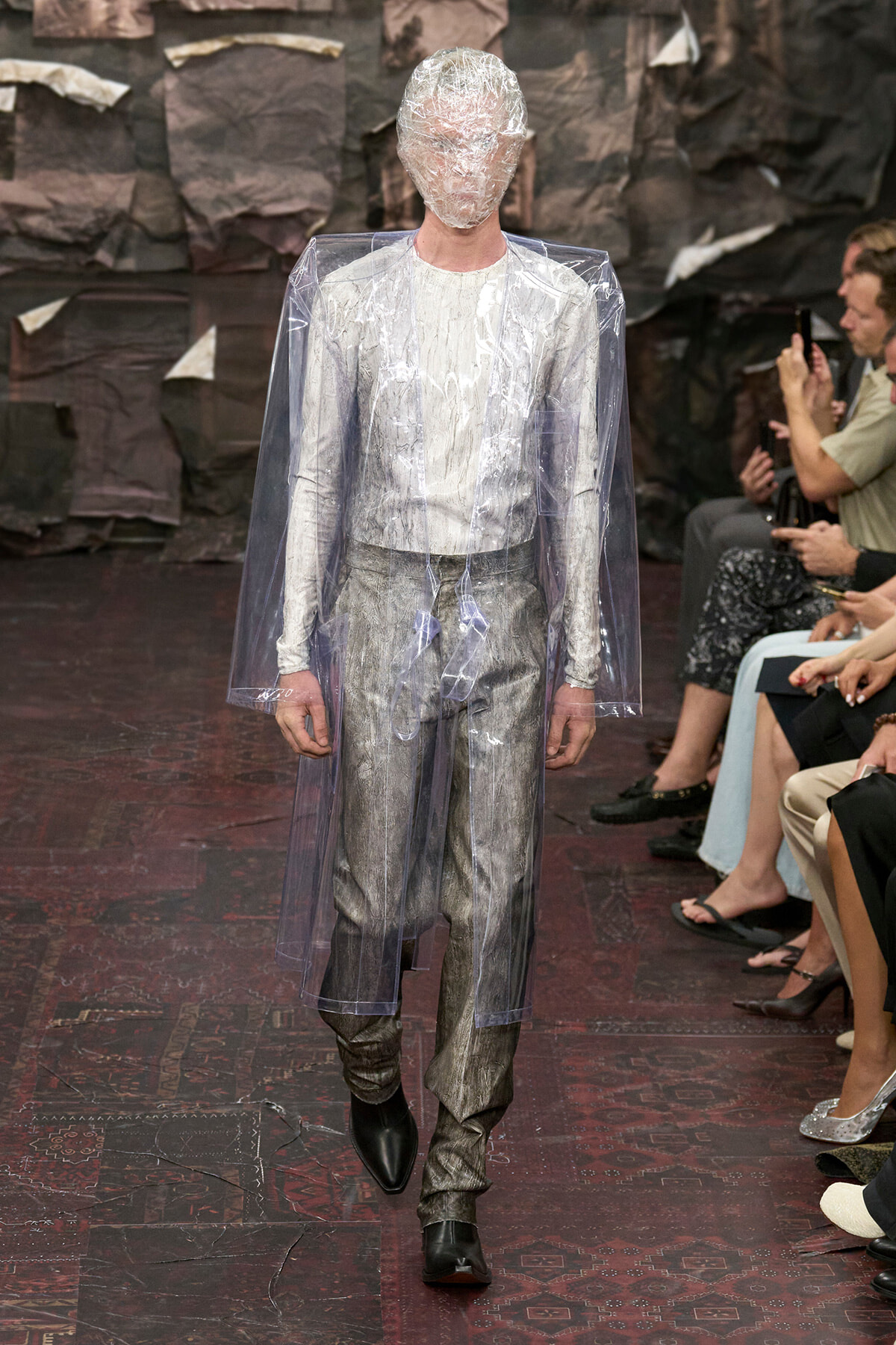 Model walks a runway in a translucent plastic cape and head covering over a metallic silver suit, with audience watching on the side.