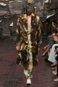Model walks the runway in an abstract camouflage suit and oversized coat, face obscured by a brown sculpted mask headpiece, backstage-like dark backdrop.