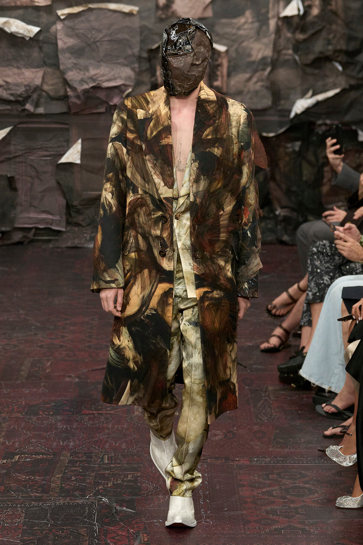 Model walks the runway in an abstract camouflage suit and oversized coat, face obscured by a brown sculpted mask headpiece, backstage-like dark backdrop.