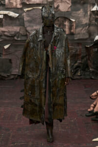 Runway model wearing a long, tattered patchwork coat with a hooded headpiece, walking on a fashion-show catwalk.