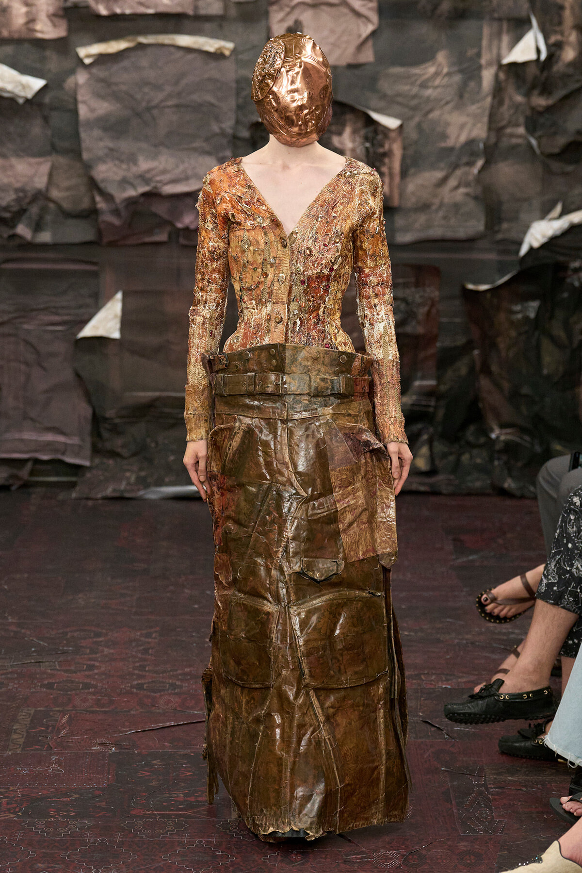 Model on a runway wearing a copper-toned, textured gown with a matching metallic hooded mask.