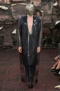 Model walking a runway in a long, transparent PVC coat over a dark outfit, with a metallic foil headpiece covering the head.