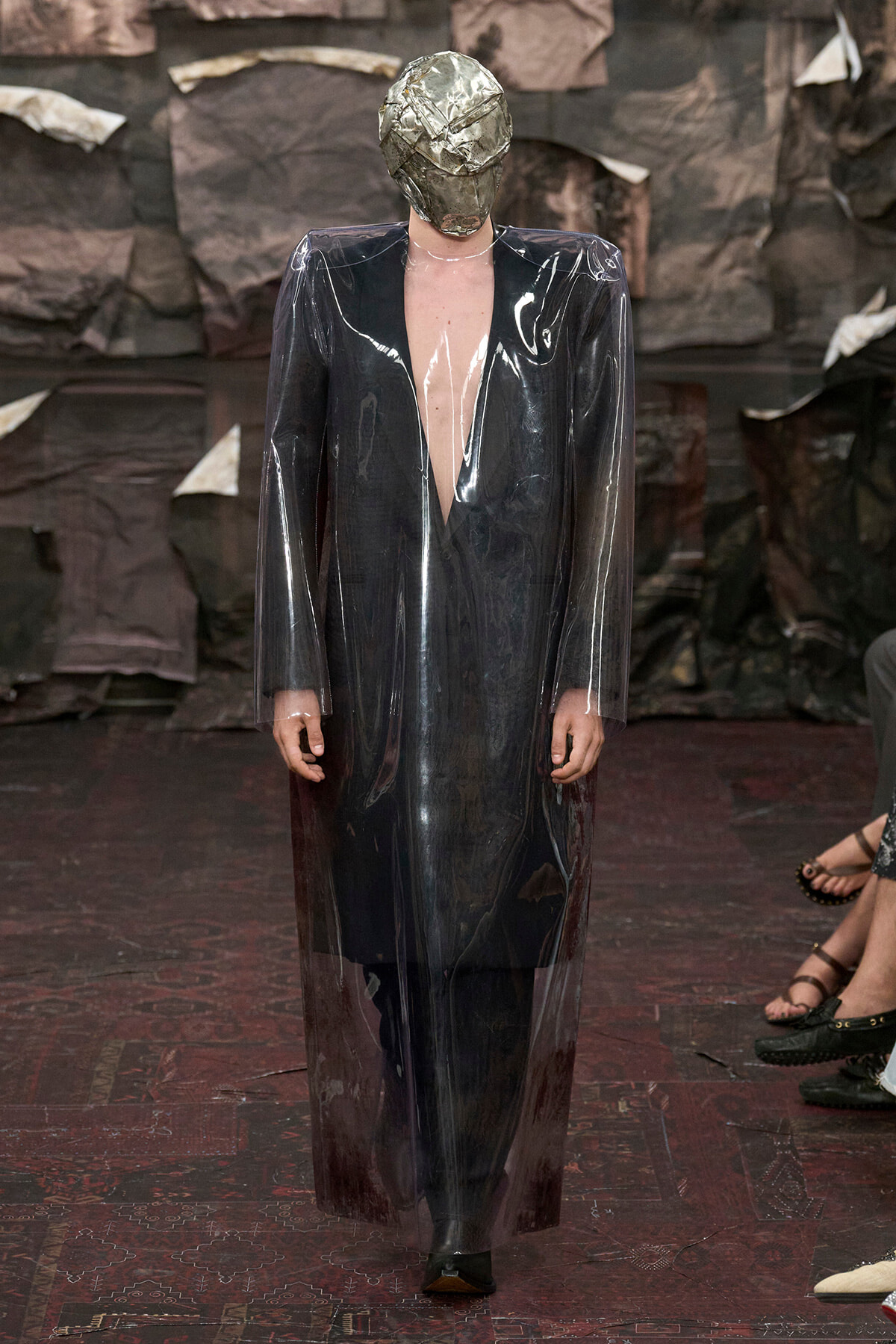 Model walking a runway in a long, transparent PVC coat over a dark outfit, with a metallic foil headpiece covering the head.
