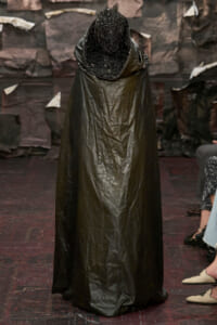 Model in a long, glossy black cape with an ornate beaded hood walking the runway at a fashion show