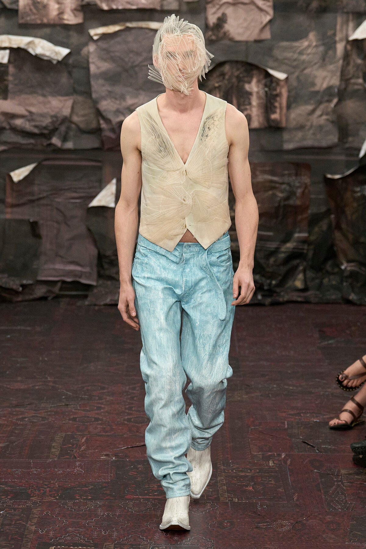 Male runway model walking the catwalk in a beige sleeveless top with leaf-like folds and light blue loose pants, with a translucent wrap over the head.