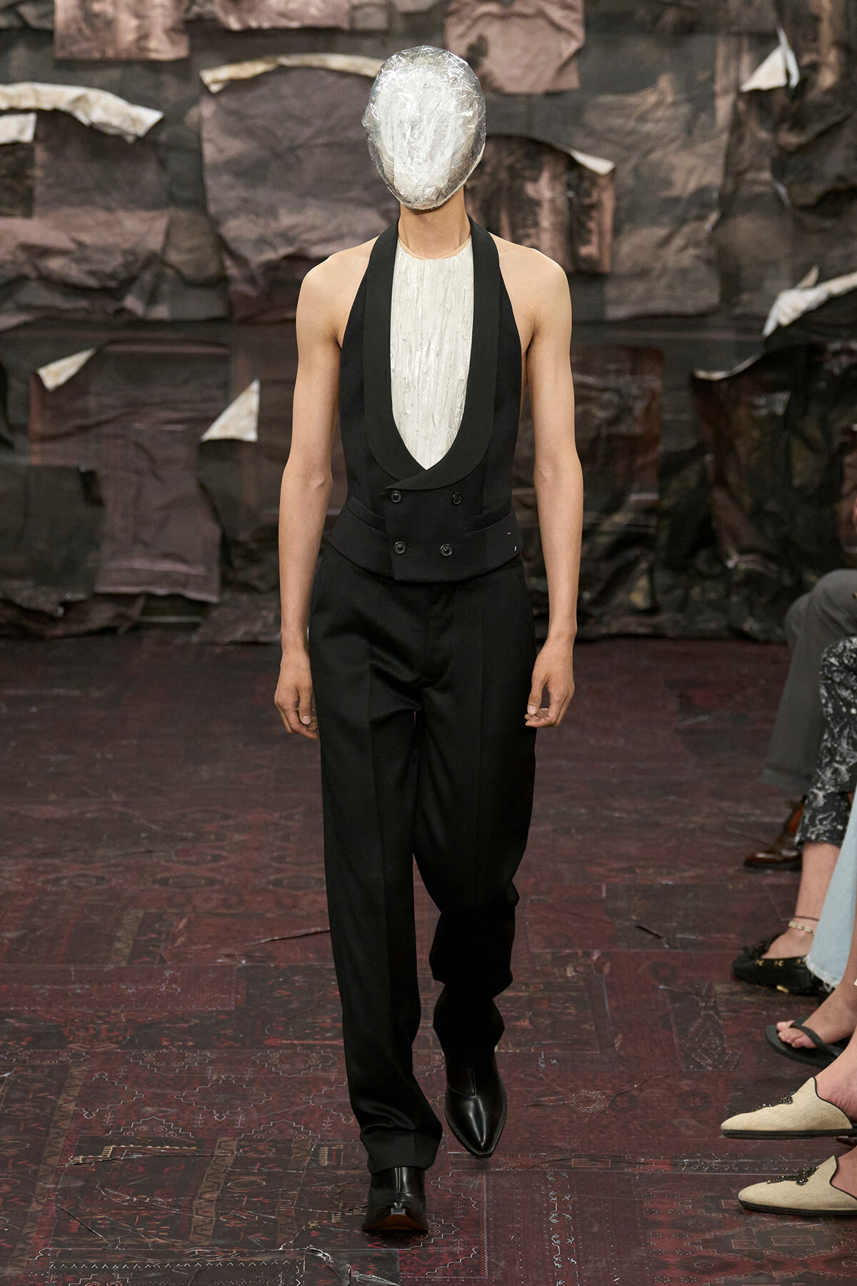 Model on a runway wearing a black vest over a white textured top with wide black trousers and pointed boots; head wrapped in a metallic foil mask.