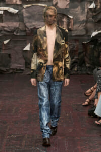 Male model on a runway wearing an open camouflage blazer, bare chest, acid-wash jeans, and brown boots.