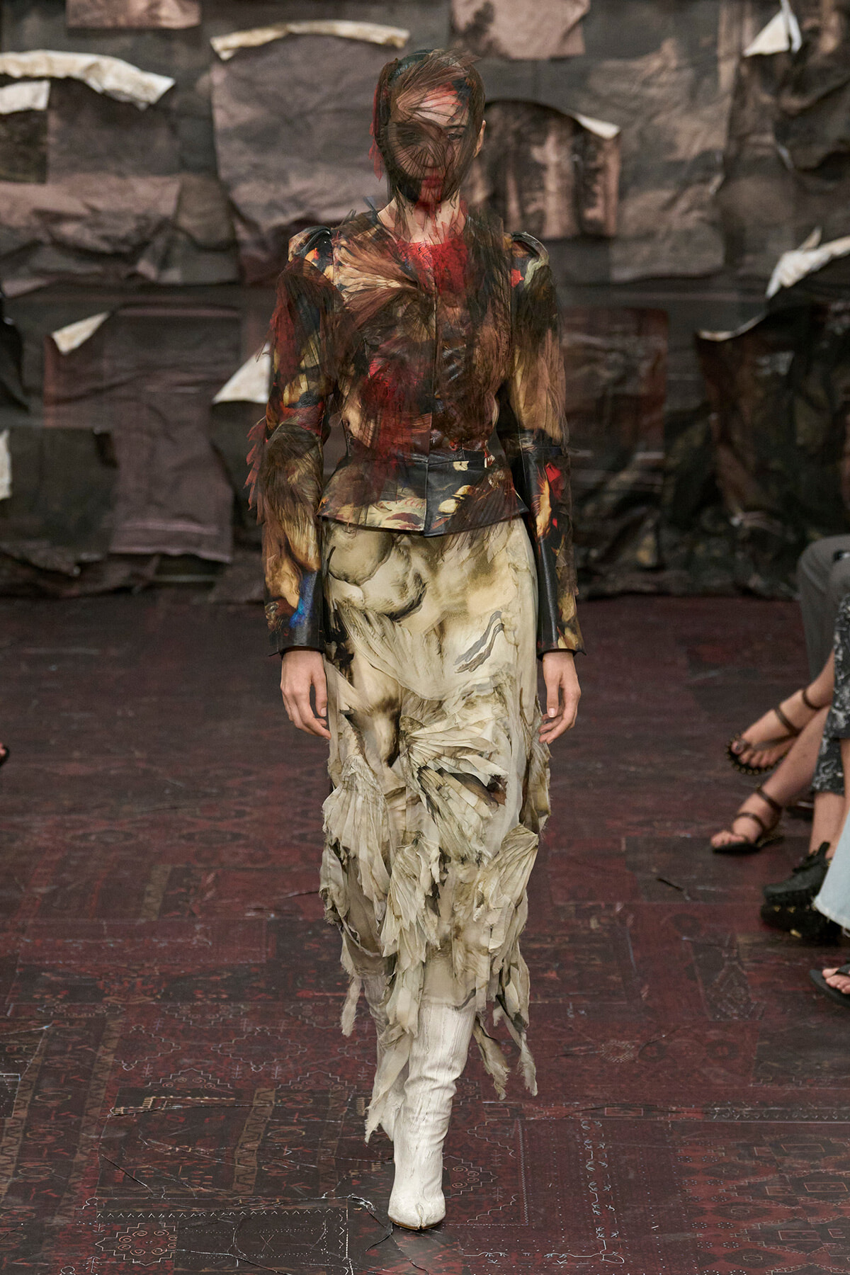 Runway model in a distressed multicolor top and a long, frayed beige skirt with white boots, walking down a dark, textured catwalk.