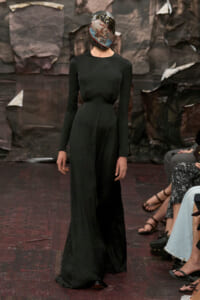 Model walks the runway in a black, long-sleeve, floor-length gown with a glossy, collage-covered headpiece obscuring the face.