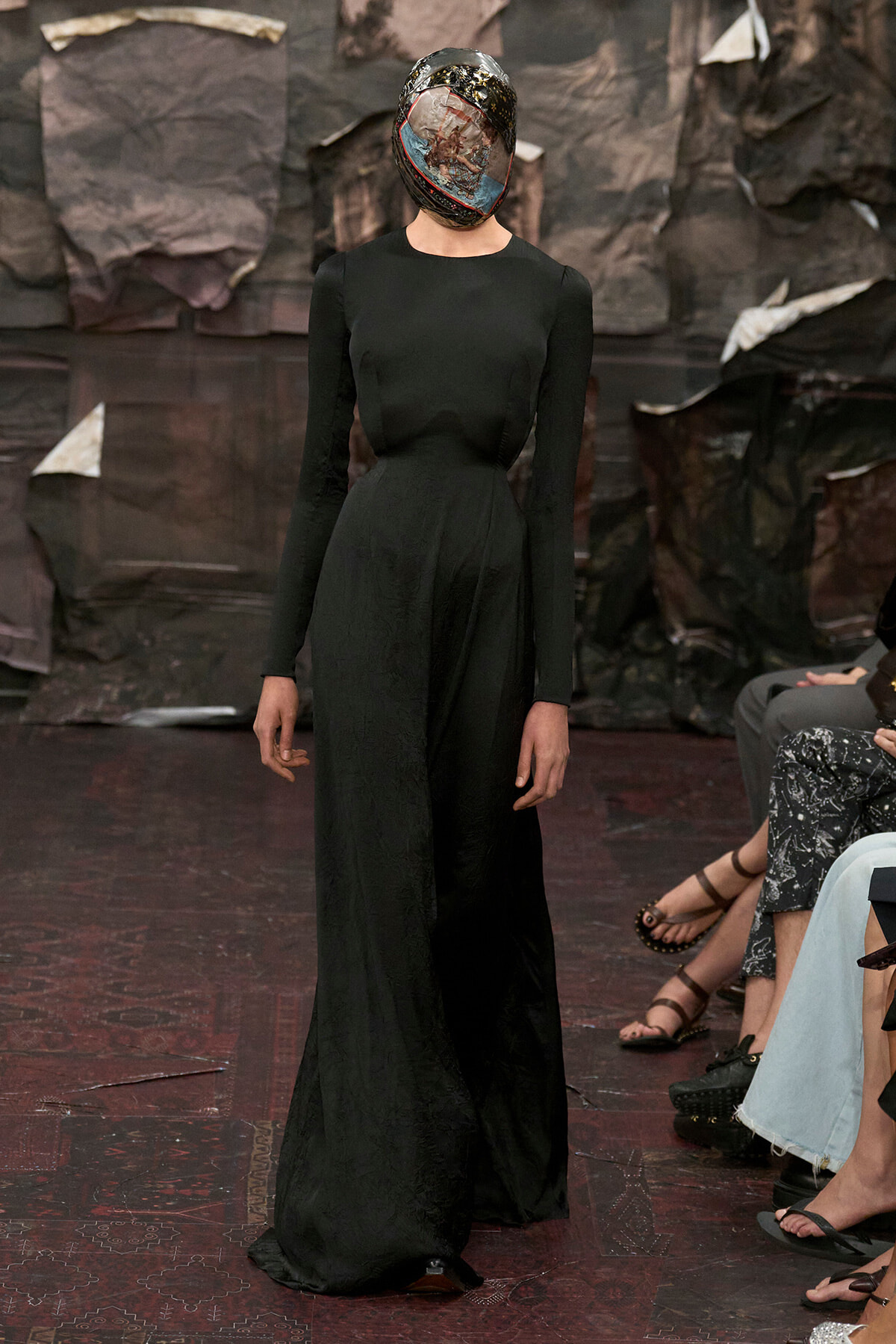Model walks the runway in a black, long-sleeve, floor-length gown with a glossy, collage-covered headpiece obscuring the face.