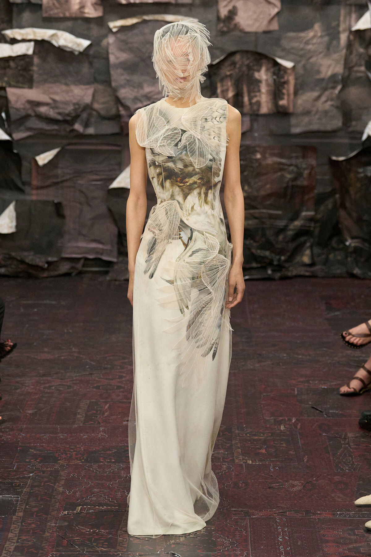 Runway model wearing a long white gown with translucent leaf appliqués and a sheer headpiece.