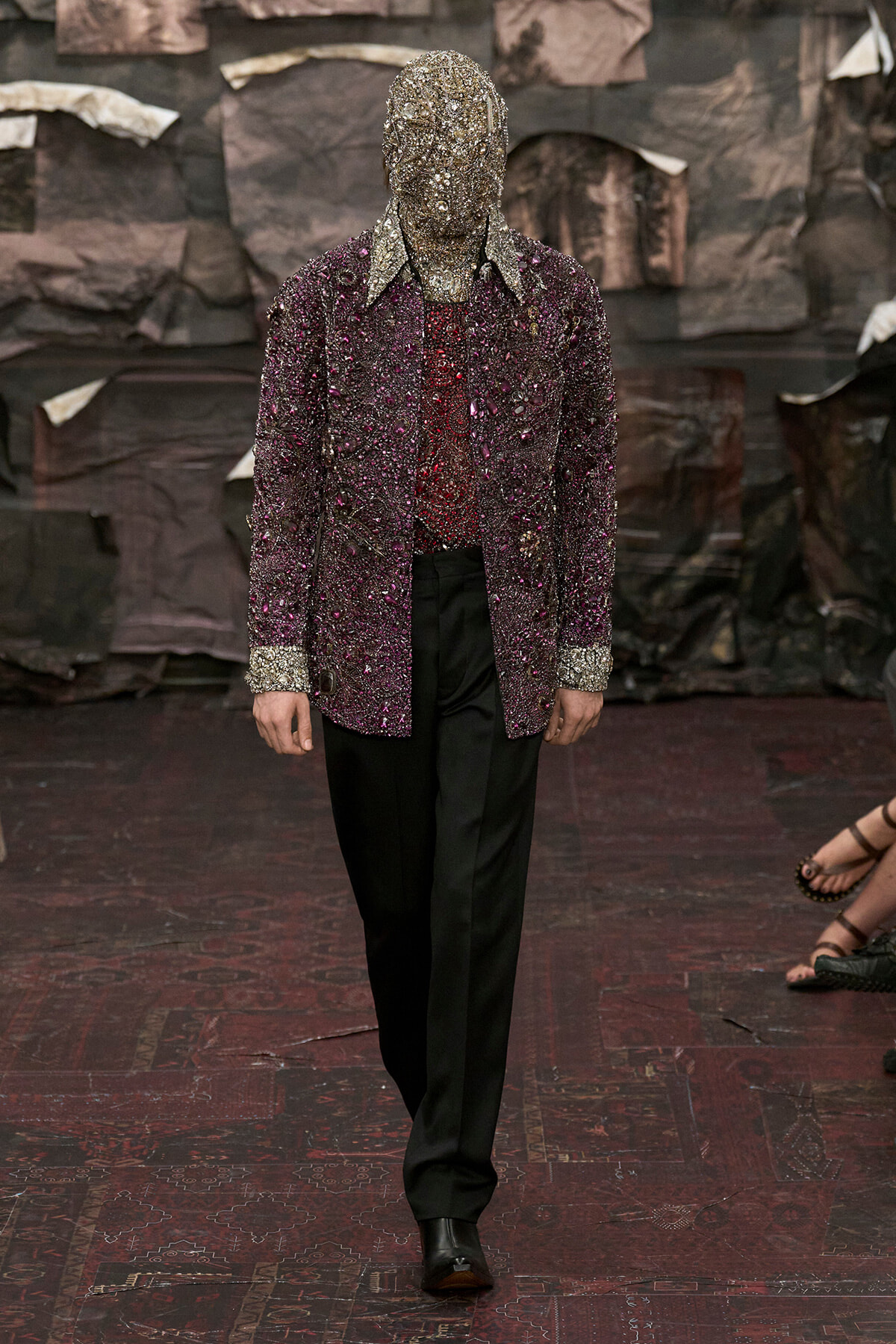Model wearing a densely beaded purple jacket with matching jeweled headpiece and black trousers on a runway.