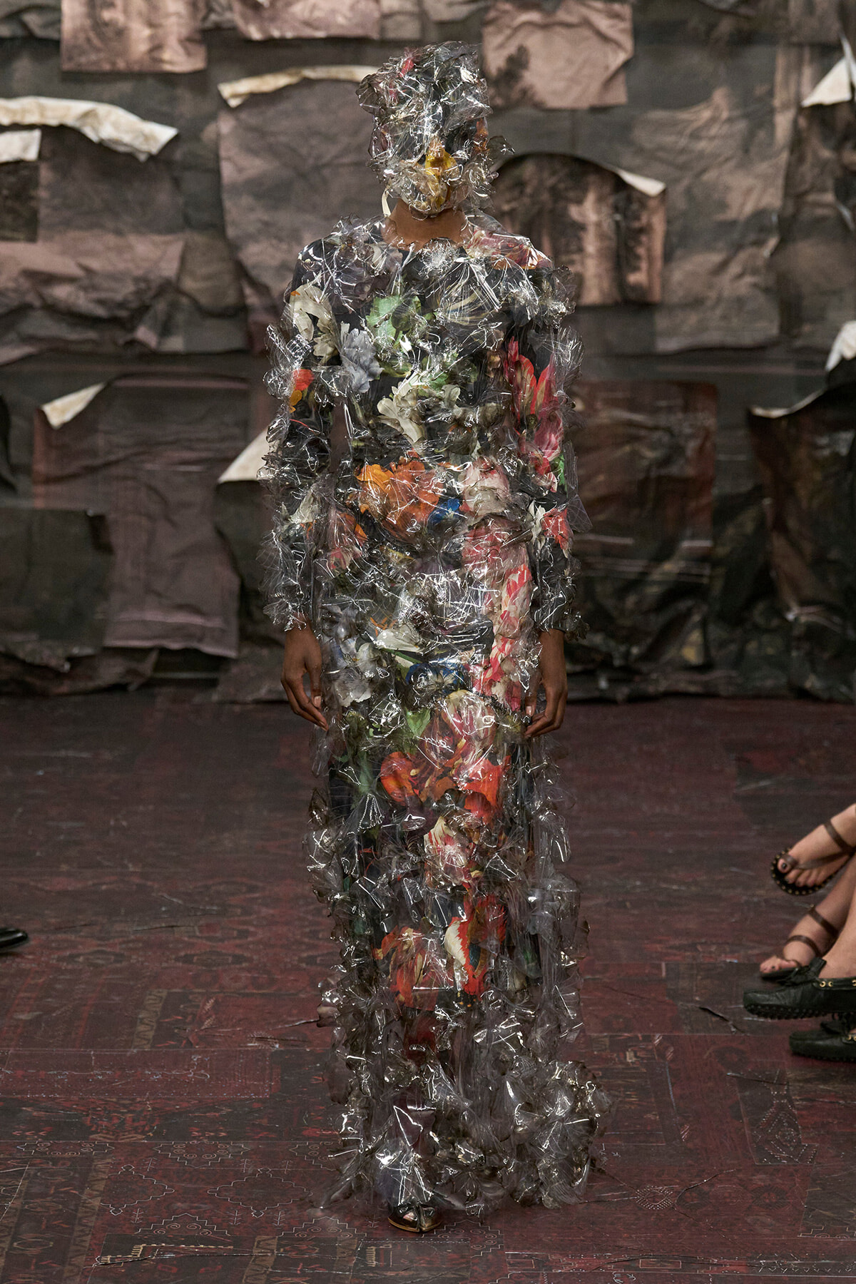 Person wearing a full-length dress and headpiece made from crumpled clear plastic wrap and assorted trash bags.
