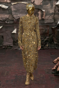 Model on a runway wearing a full-length gold dress covered in coins and a metallic gold headpiece that covers the face.