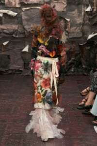Model on a fashion-show runway wearing a long floral print dress with a cream sash and sheer tulle accents.