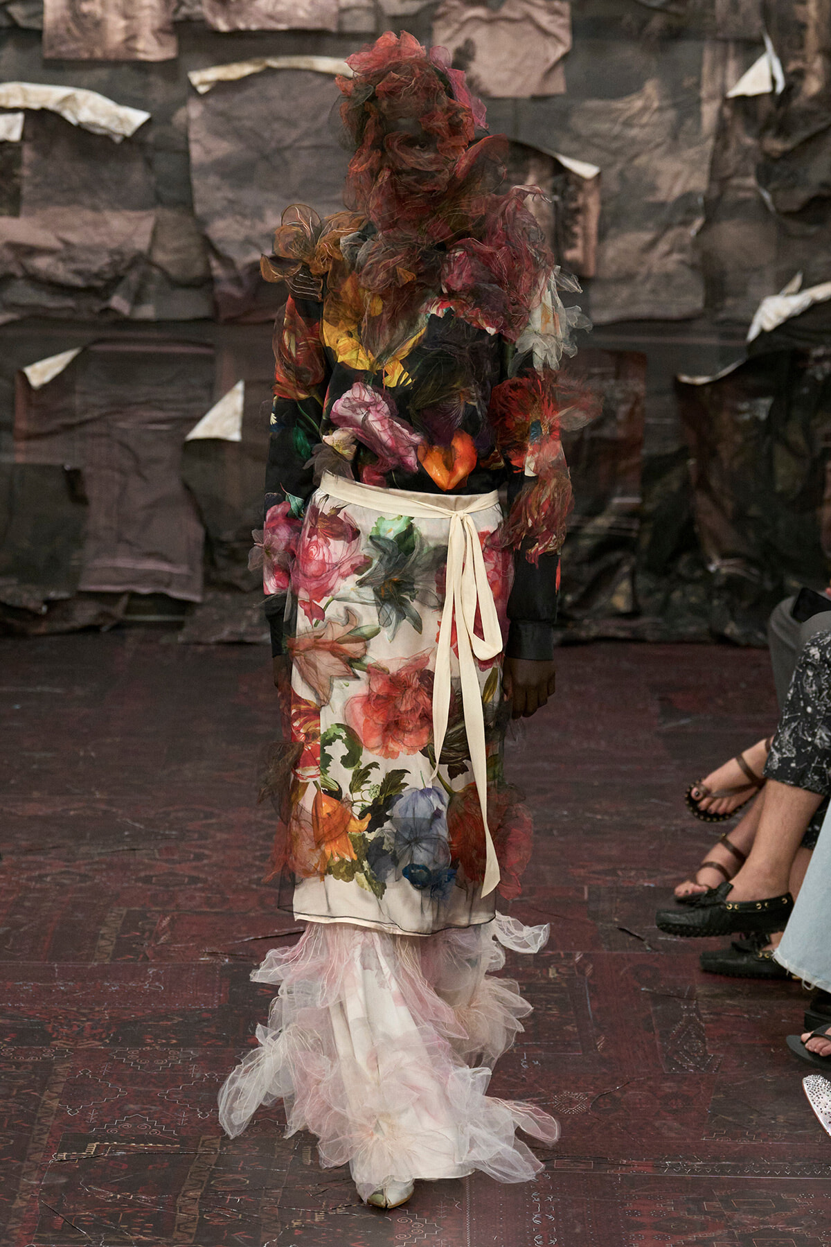 Model on a fashion-show runway wearing a long floral print dress with a cream sash and sheer tulle accents.
