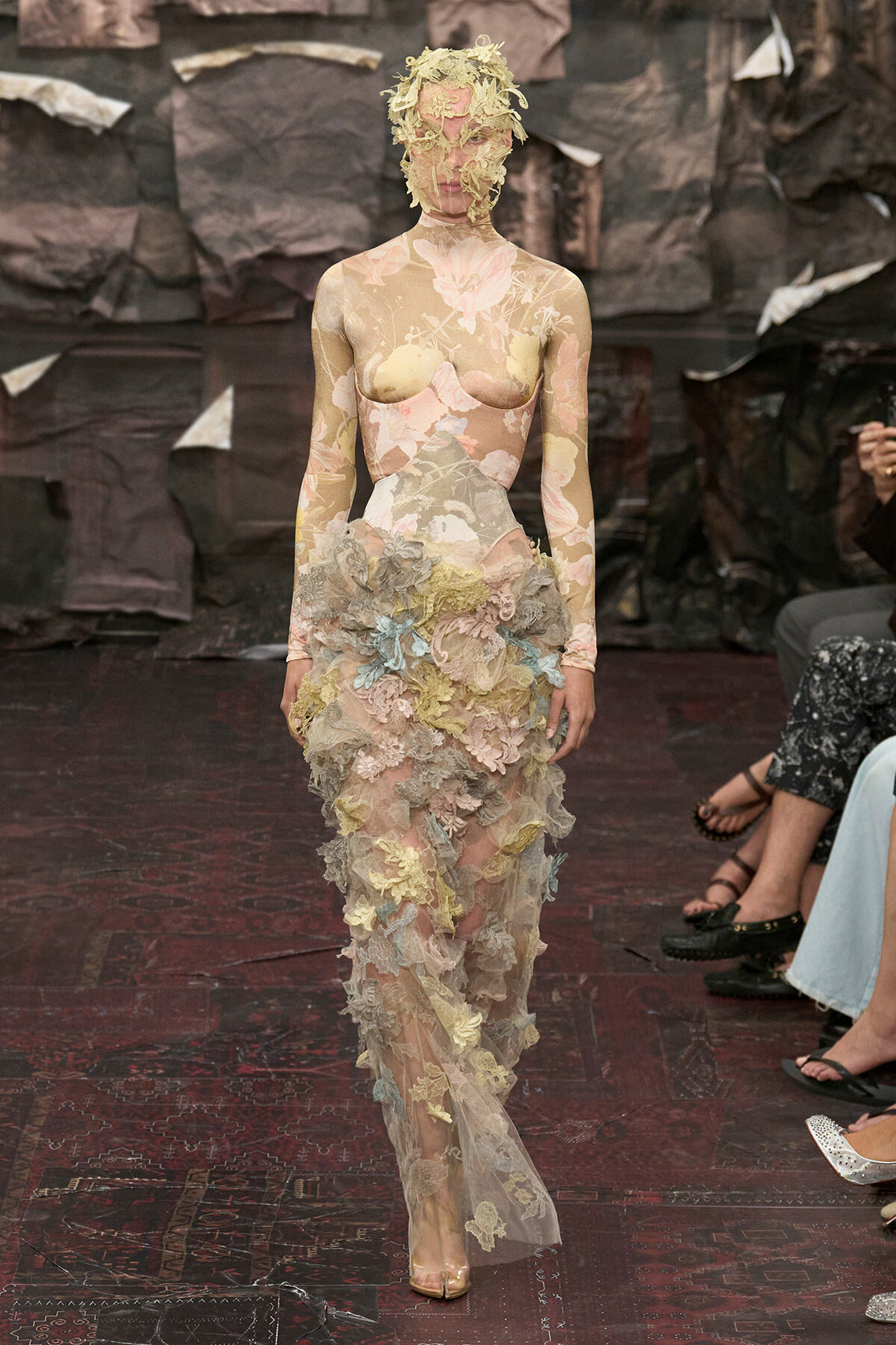 Model walking the runway in a sheer pastel floral lace gown with layered appliqués and a lace headpiece.