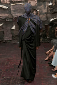 Model walks away from the camera in a long black dress with a blue patterned scarf wrapped around the head and shoulders, back view on a fashion runway.