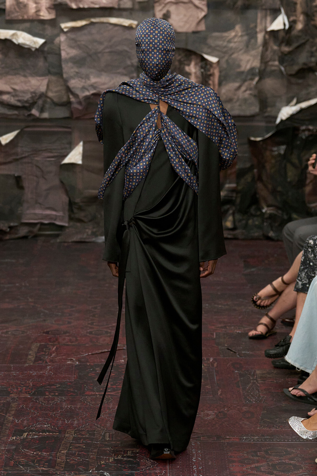 Model walks away from the camera in a long black dress with a blue patterned scarf wrapped around the head and shoulders, back view on a fashion runway.
