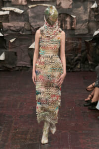 Model on a runway wearing a long beige dress made of knitted beadwork in multicolored patterns, with a metallic headpiece obscuring the face.