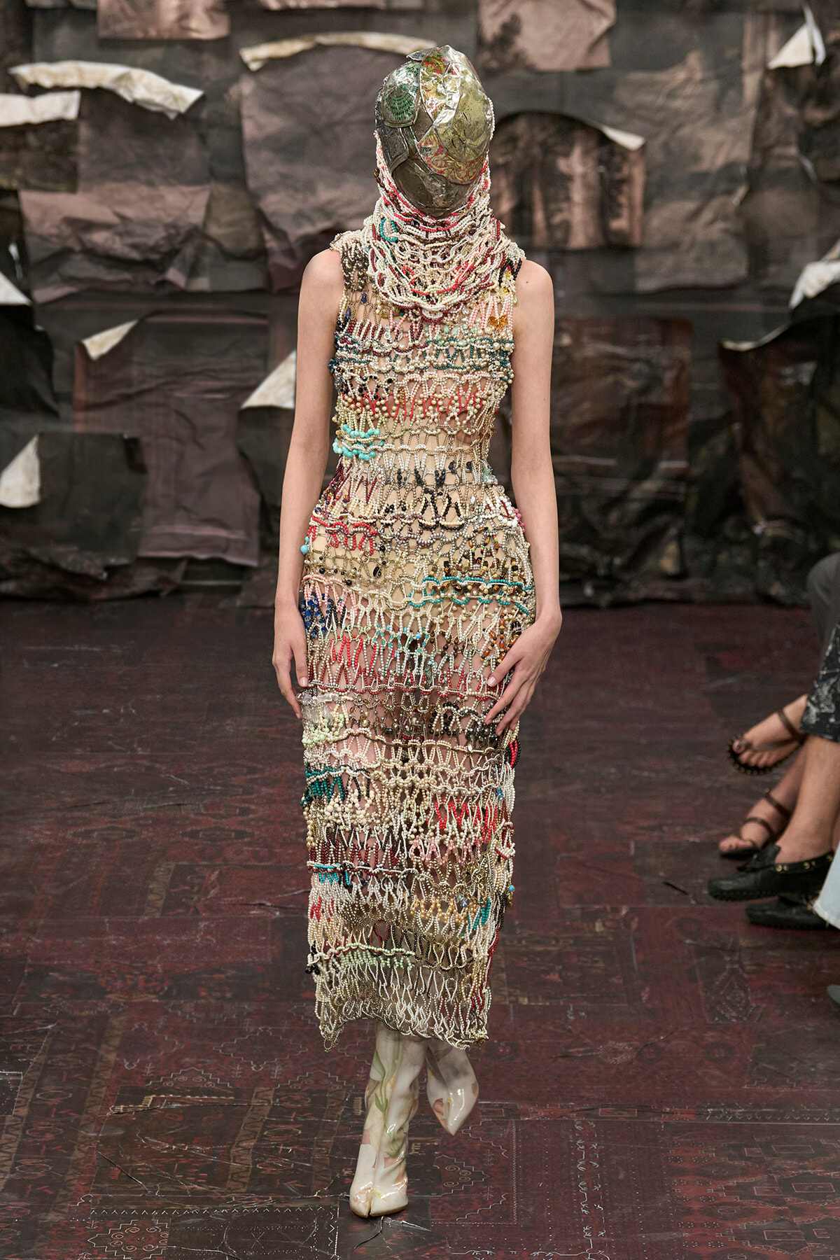 Model on a runway wearing a long beige dress made of knitted beadwork in multicolored patterns, with a metallic headpiece obscuring the face.