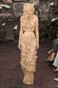 Model wearing a sheer nude gown with large beige floral appliqués and a matching floral headpiece on a runway.