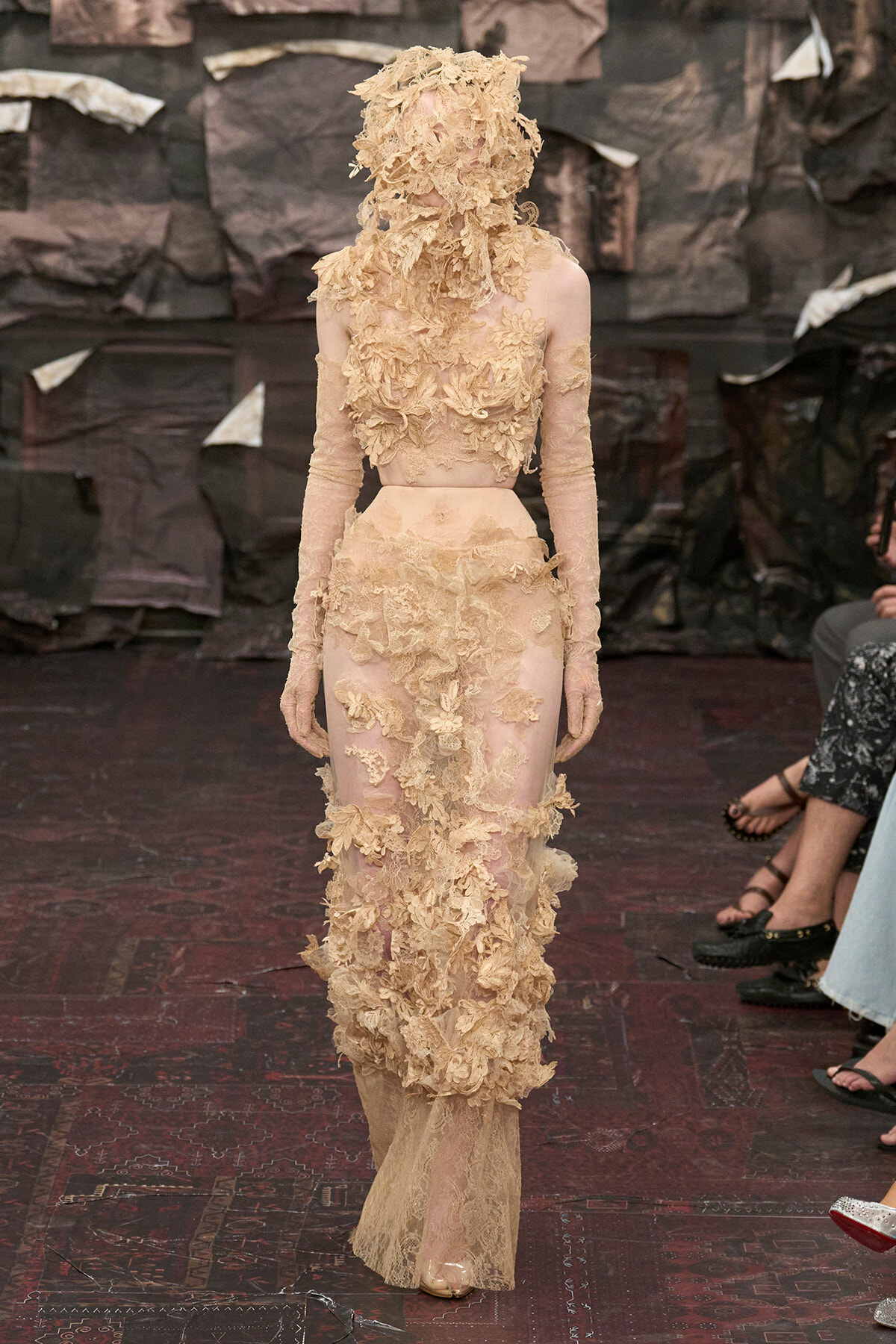 Model wearing a sheer nude gown with large beige floral appliqués and a matching floral headpiece on a runway.
