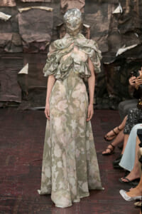 Model walks the runway in a pale green floral gown with dramatic ruched shoulders and a metallic helmet-like headpiece, backstage-style backdrop behind.