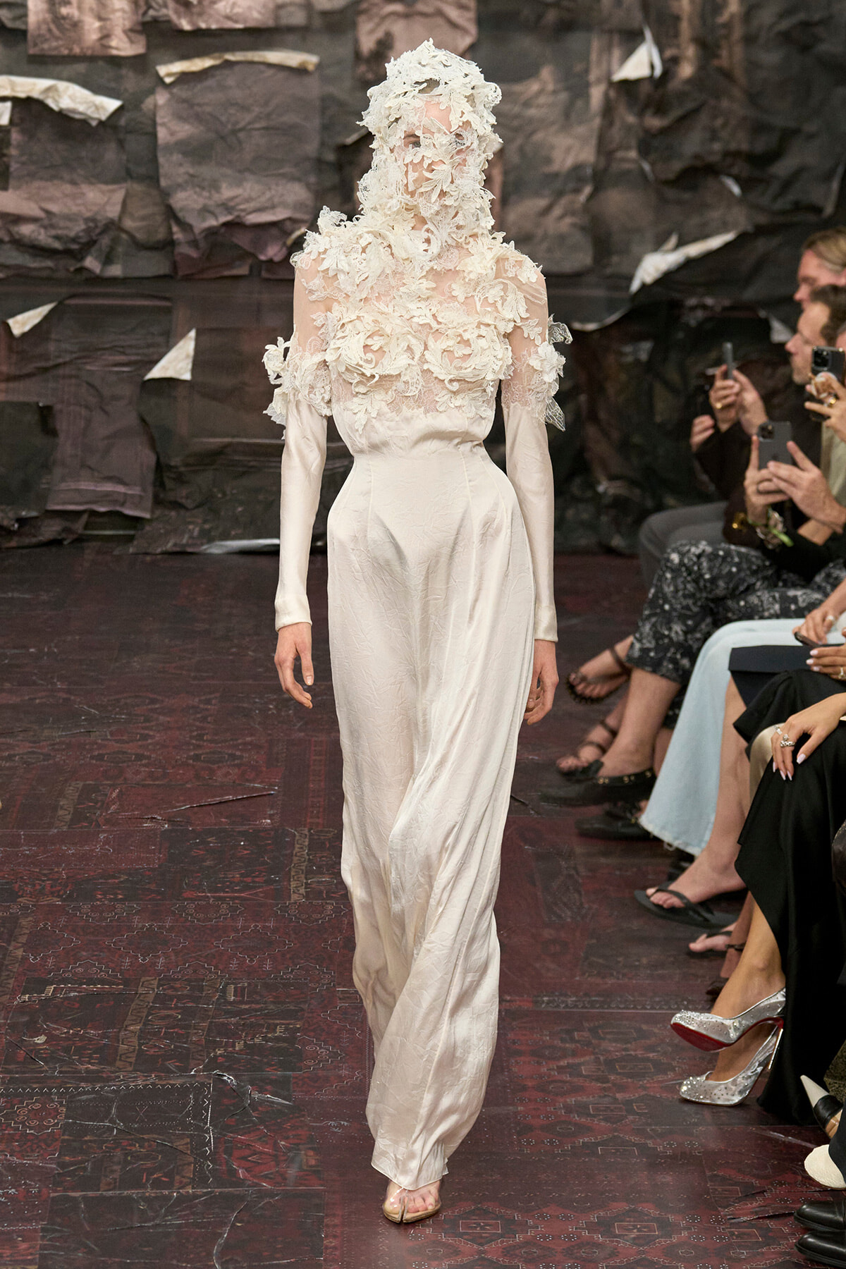 Model on a runway wearing an ivory gown with a lace floral applique bodice and matching headpiece and veil-like covering.