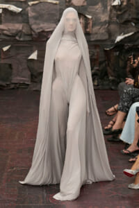 Model on a runway wearing a pale gray full-body gown with a long, sheer cape draped over the head and shoulders.