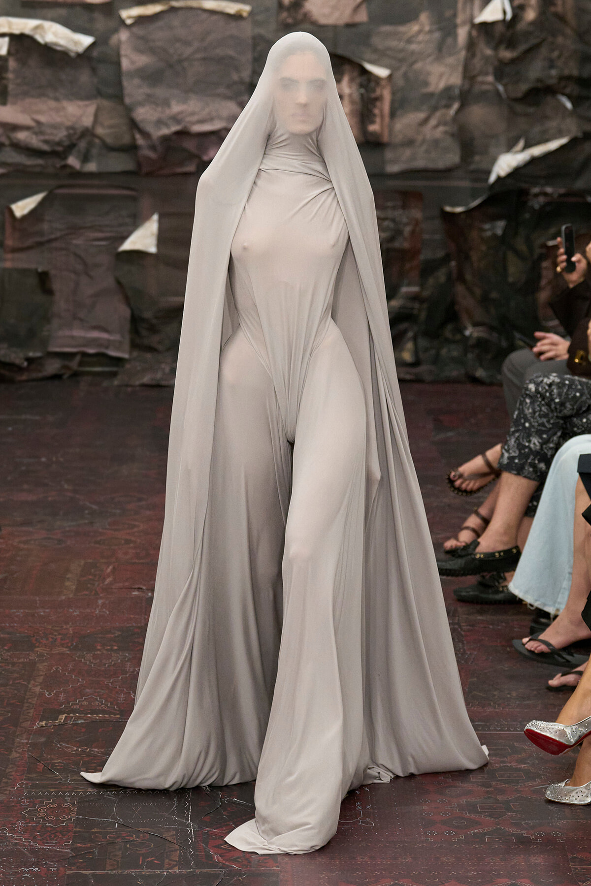 Model on a runway wearing a pale gray full-body gown with a long, sheer cape draped over the head and shoulders.