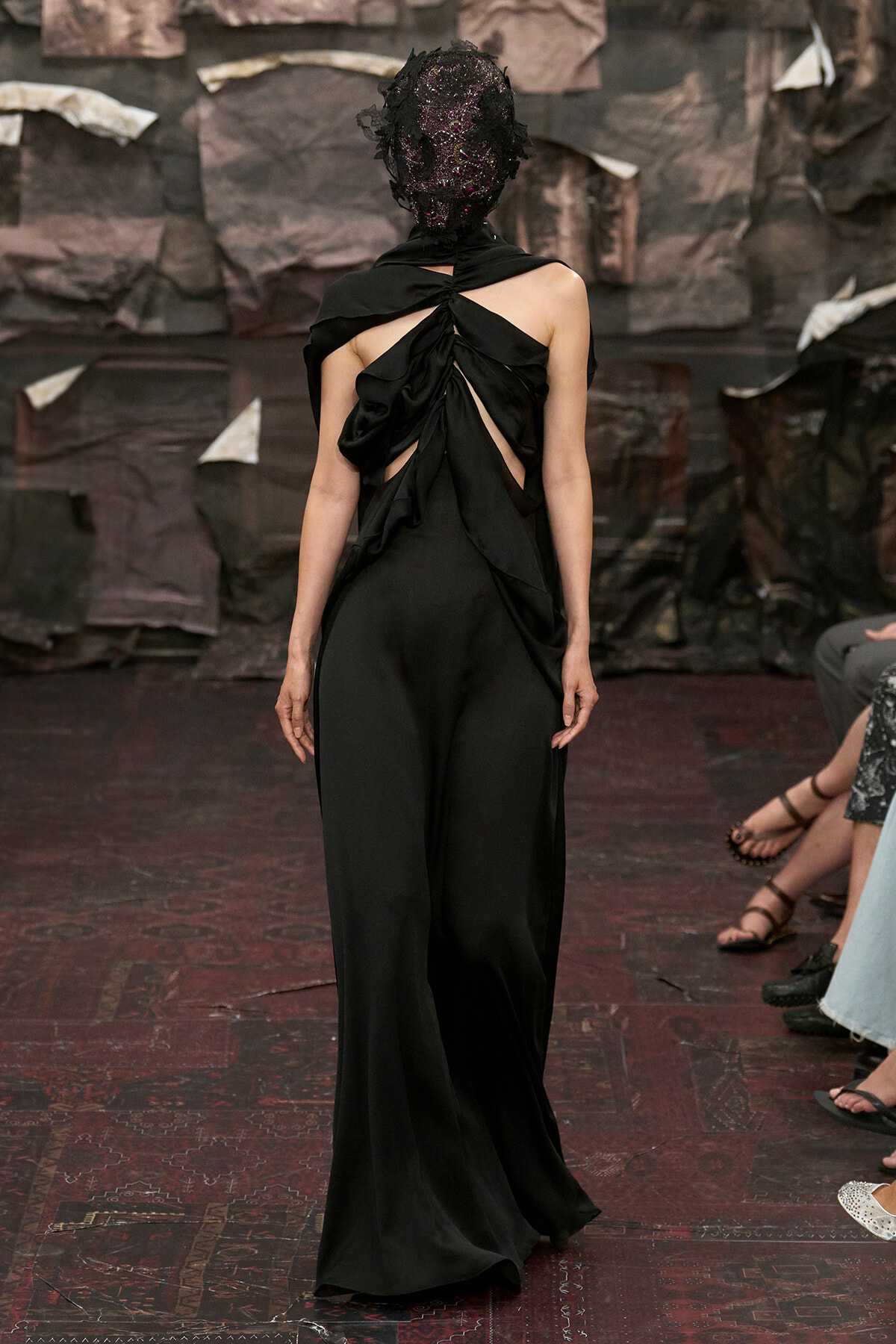 Model walks the runway in a backless black gown with twisted drape and a dark textured mask influence on the headpiece.