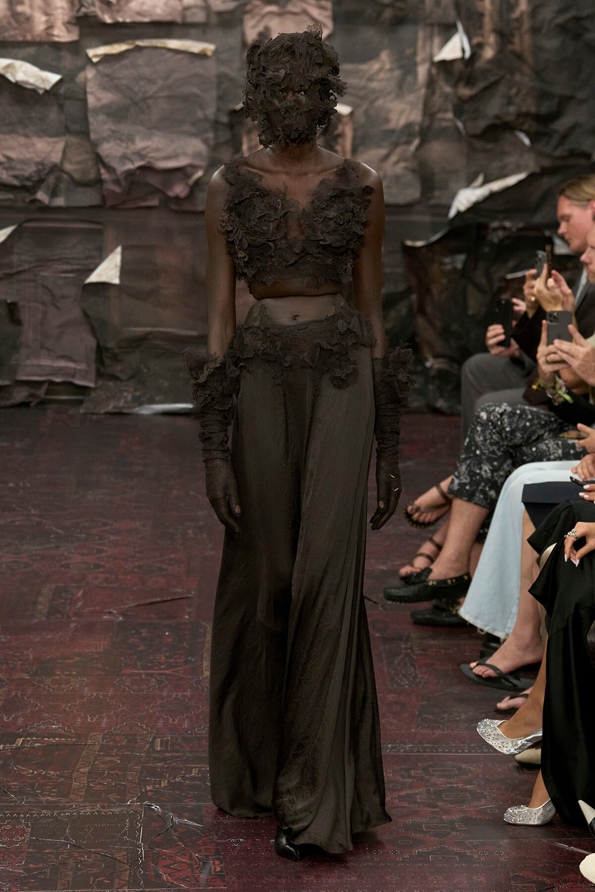 Model on a runway wearing a sheer black gown with floral appliqués and a textured hooded headpiece; audience members watch and photograph on the right.