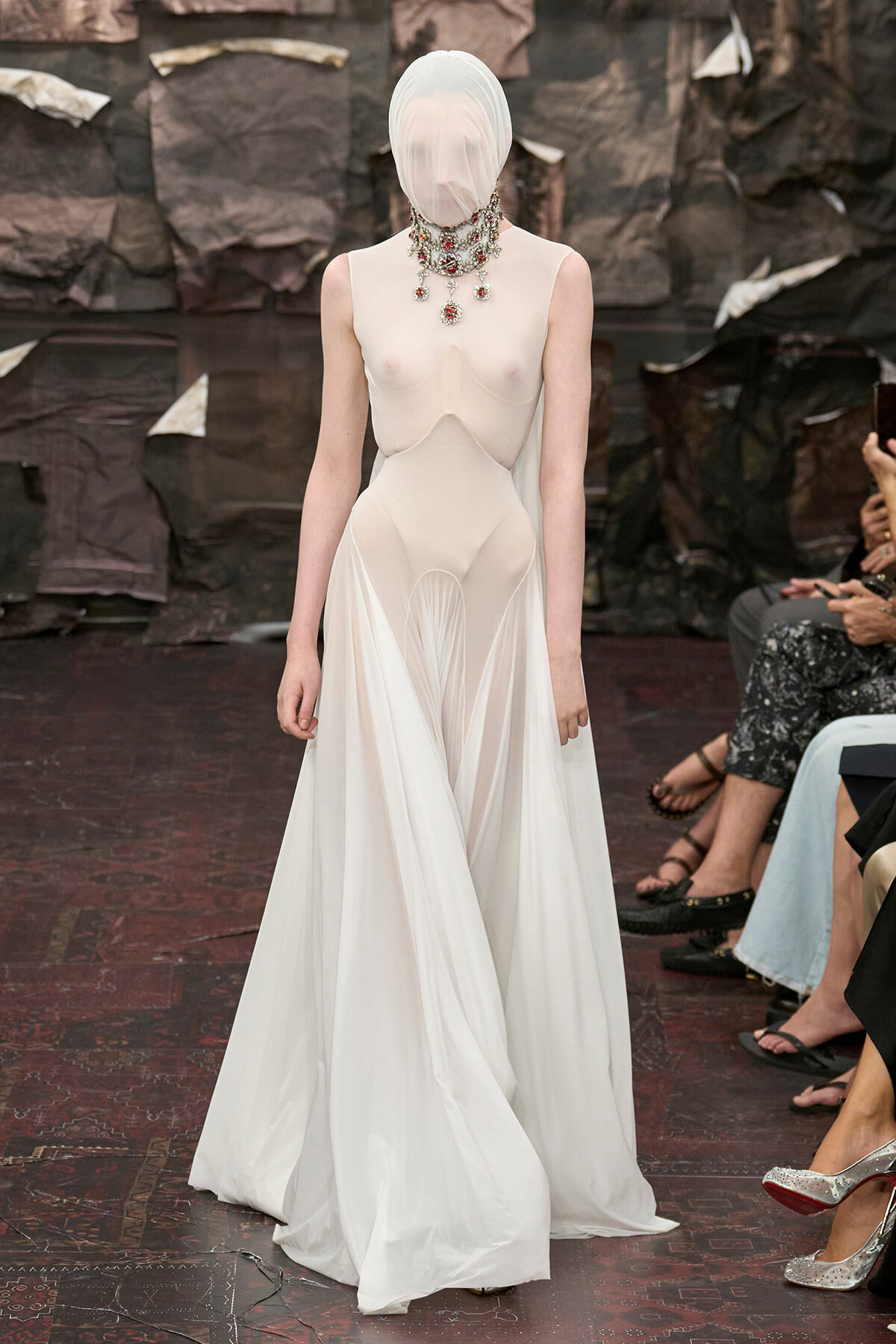 Model walking the runway in a sheer, ivory gown with a sculptural silhouette and a layered jeweled necklace; face wrapped in white fabric