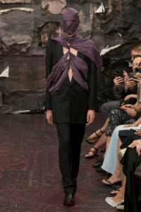 Model in a black suit with a purple dotted scarf wrapped over the head and tied at the back, creating shoulder openings.