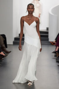 Female model walks the runway in a white, spaghetti-strap slip dress with layered panels, wearing dangling earrings and a necklace.