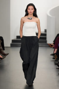 Female model on a runway wearing a white strapless ribbed top and oversized black trousers, accessorized with a chunky silver necklace.